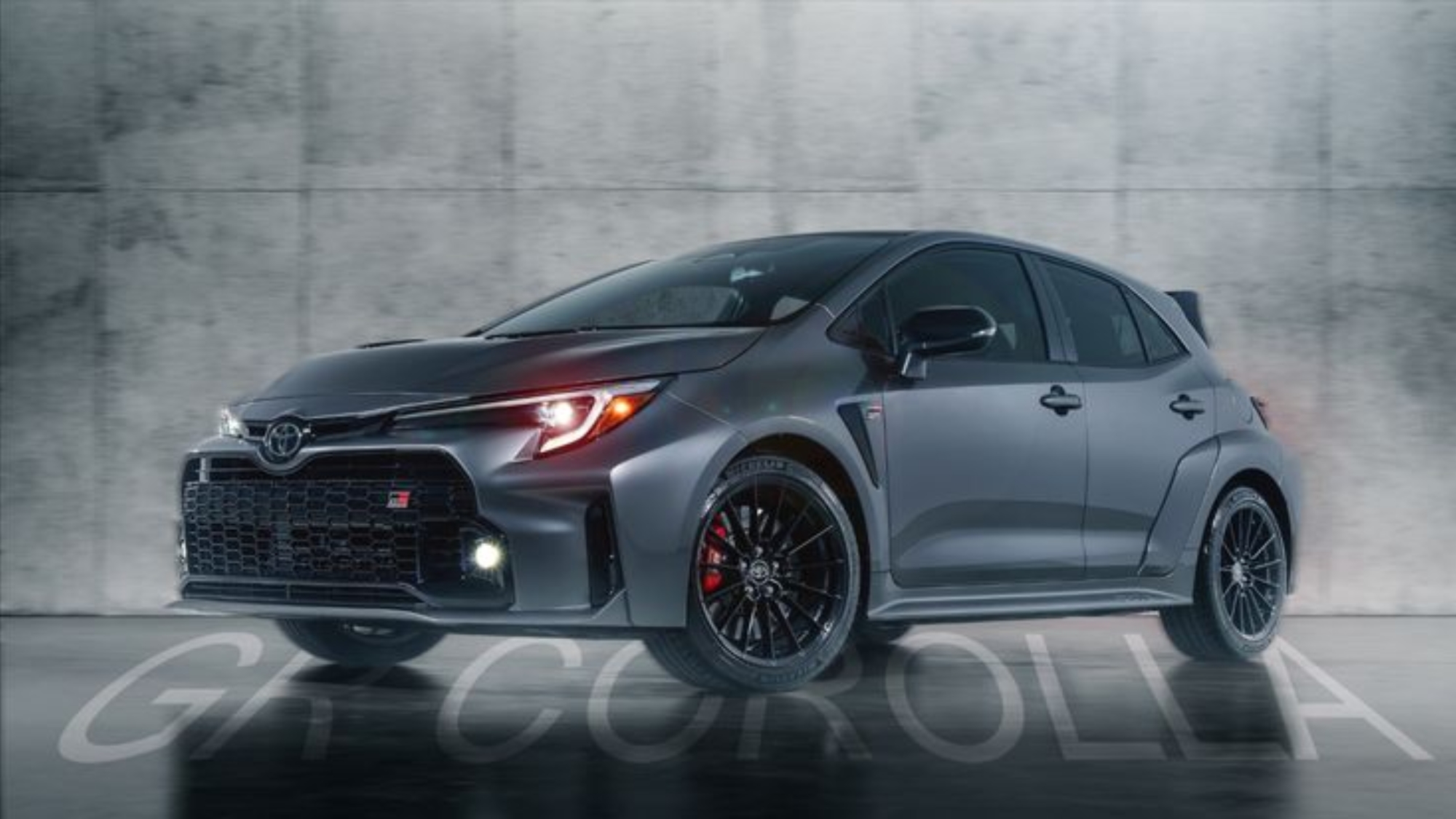 Toyota GR Corolla in metallic gray with black alloy wheels, aggressive grille, and sporty design, captured against a minimalist concrete background.